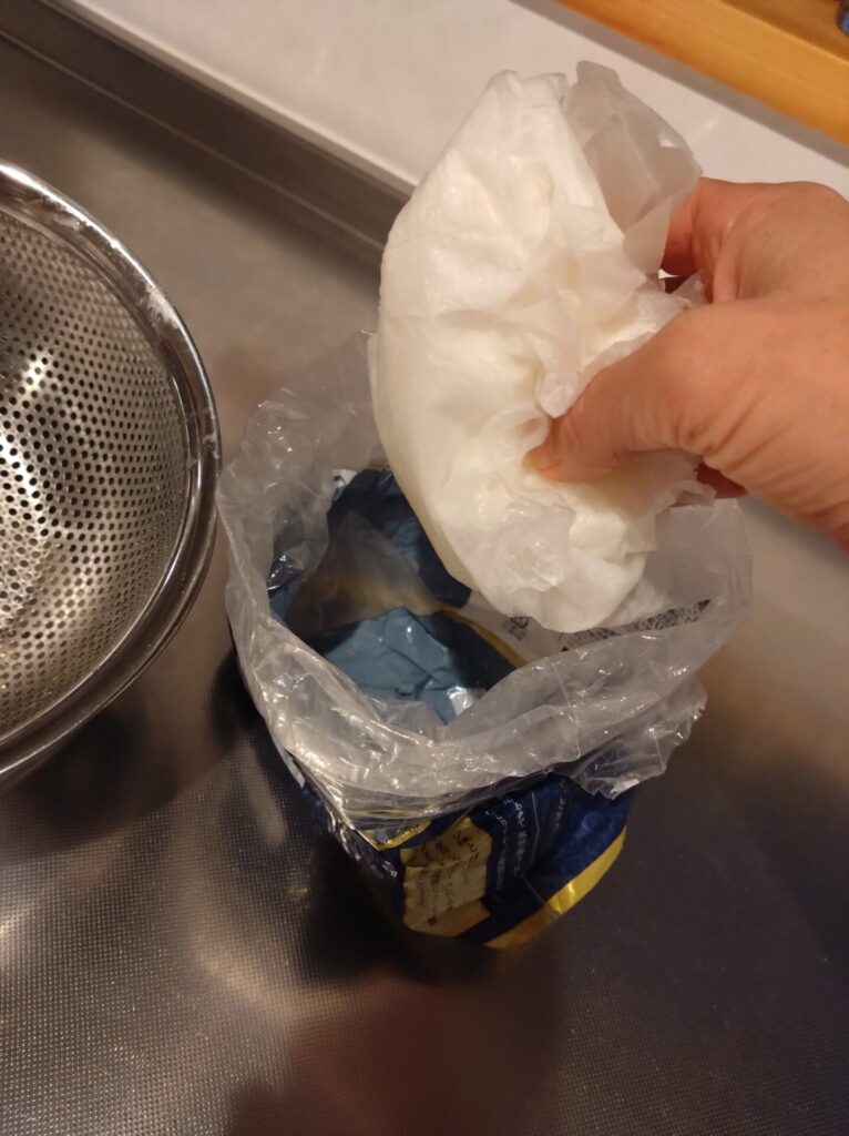 ヨーグルトを水切りしたザルからキッチンペーパータオルを掴みパンの空き袋に入れようとしている
