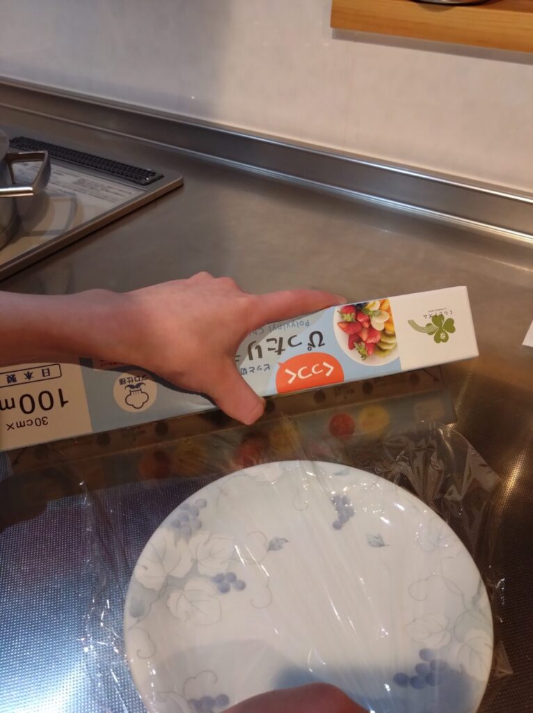 子どもが左手にラップを持ち右手でラップの端を押さえラップを切ろうとラップの箱の中心に親指を添え箱を下側へ傾けている